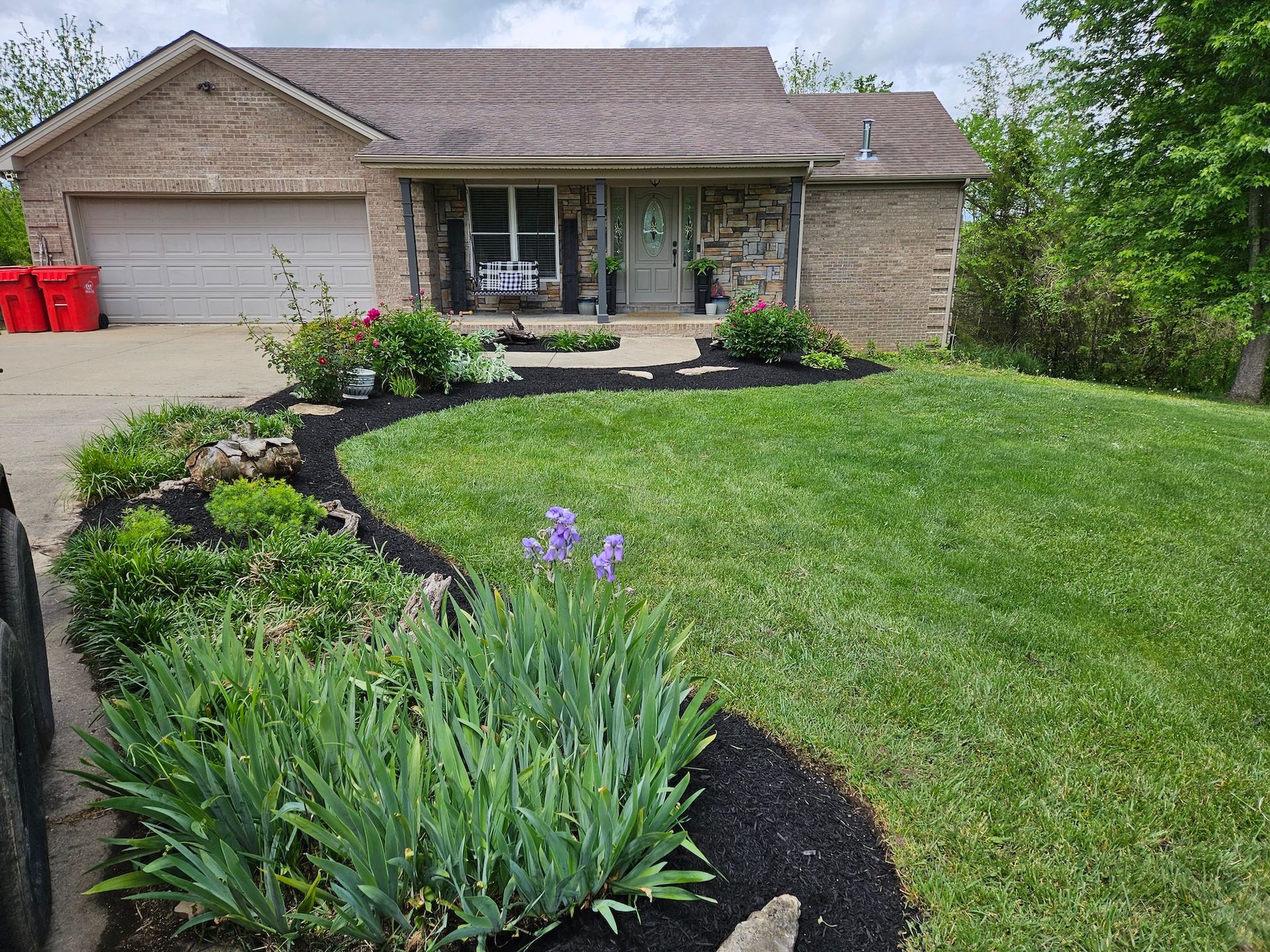 A brick house with a well-manicured lawn and flower beds, including purple irises, under a cloudy sky.