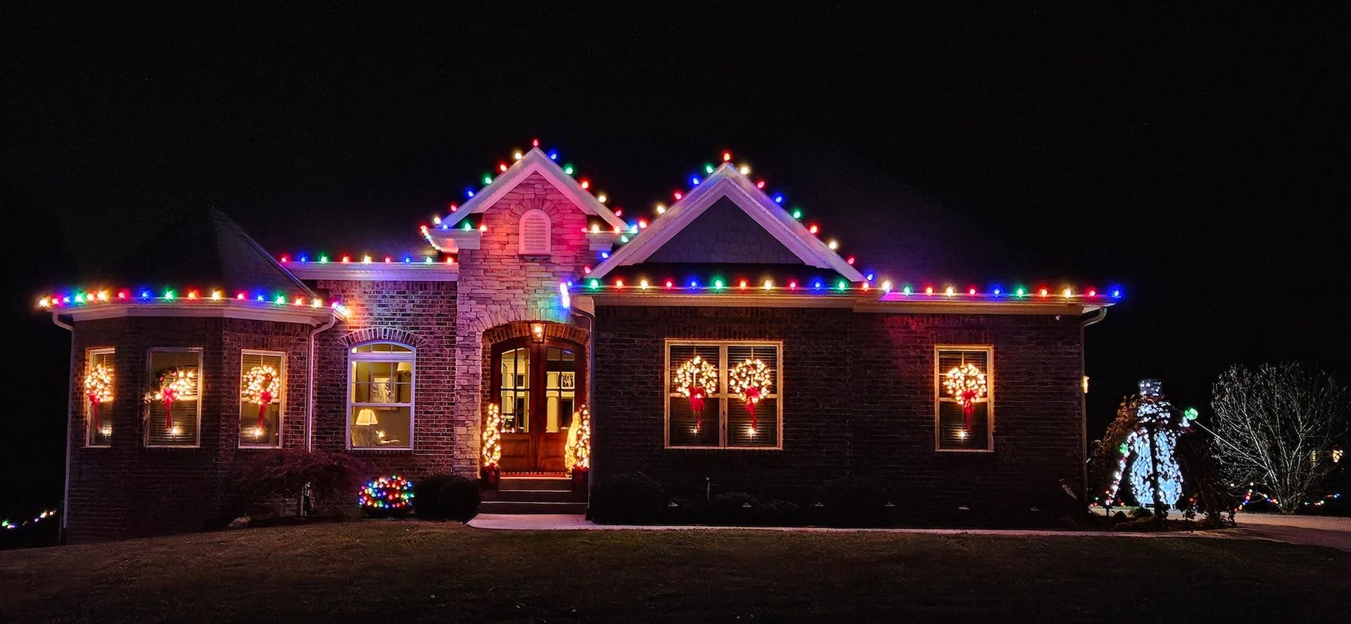A house decorated with Christmas lights, including window wreaths. A snowman decoration is in the yard.