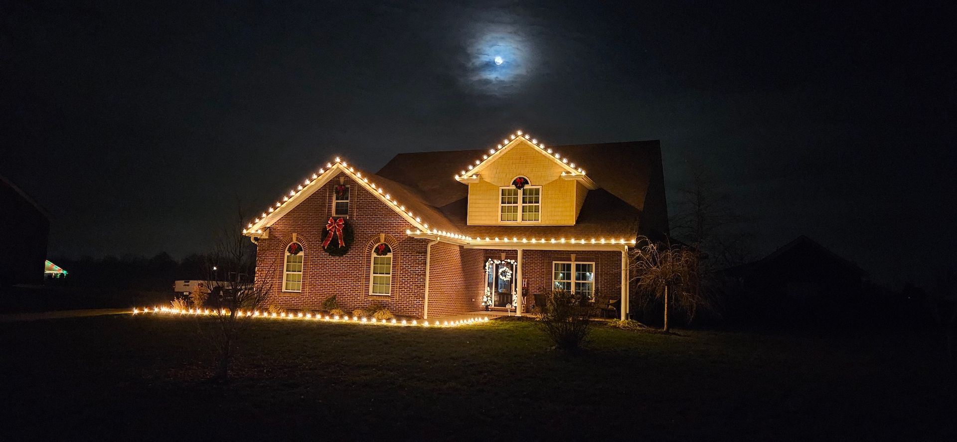 House lit with Christmas lights at night with the moon in the background.