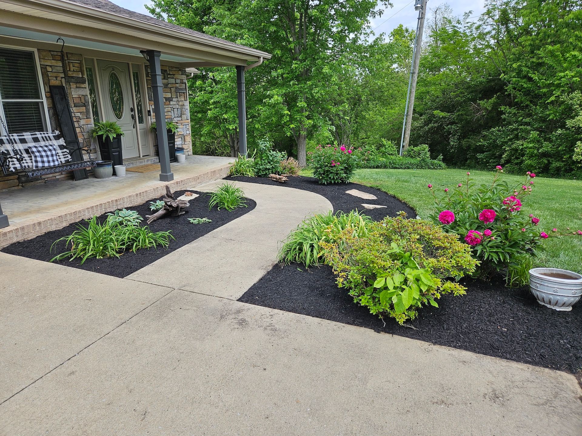 A concrete walkway curves through a landscaped yard with black mulch and flowering plants, leading to a home's porch.