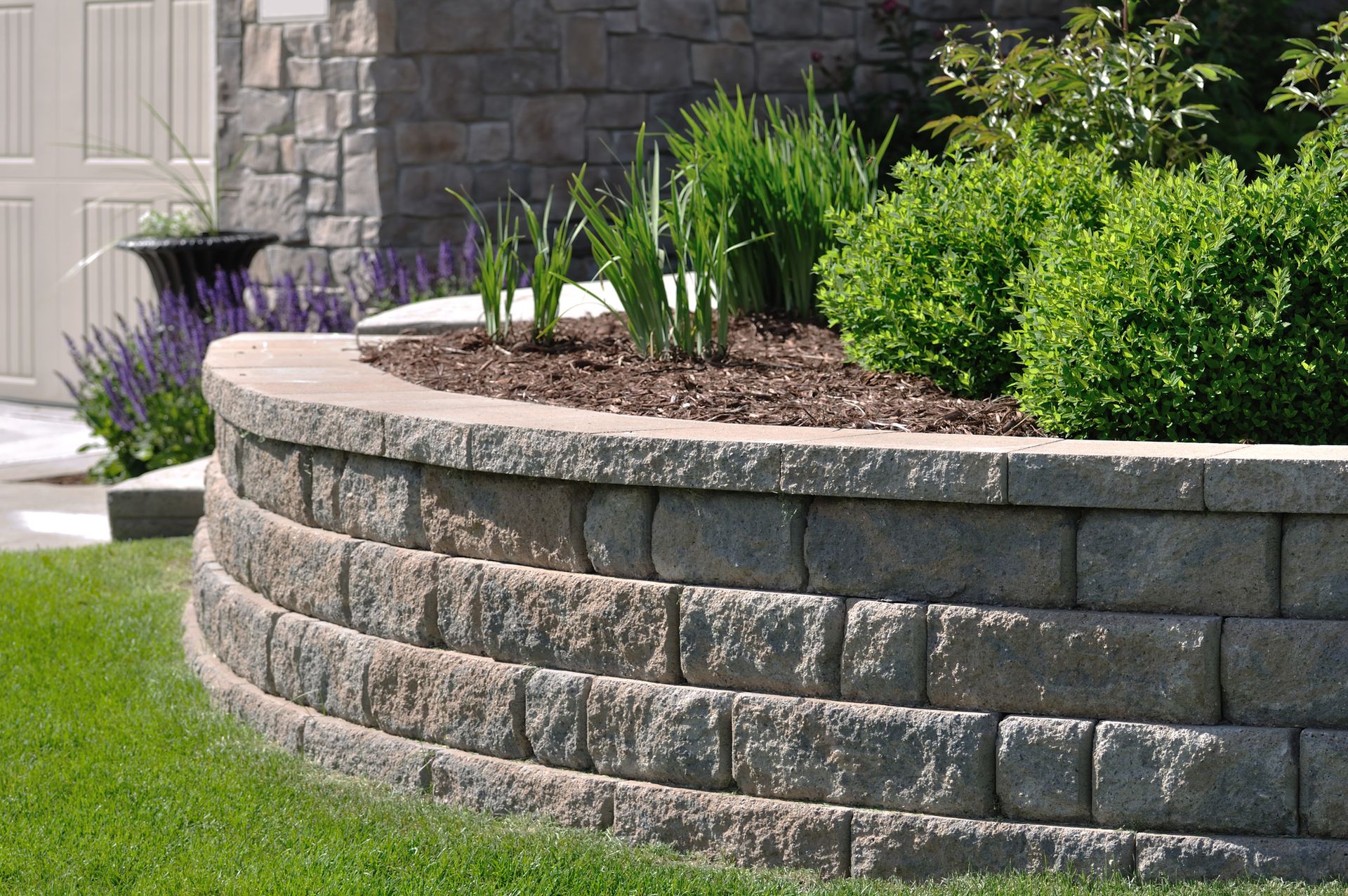 Curved retaining wall made of gray stone blocks with a garden bed featuring plants and mulch.