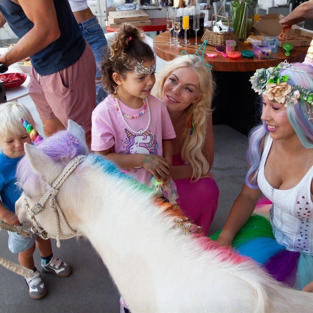 Children and adults pet a white pony at a colorful party with flower crowns and bright costumes.