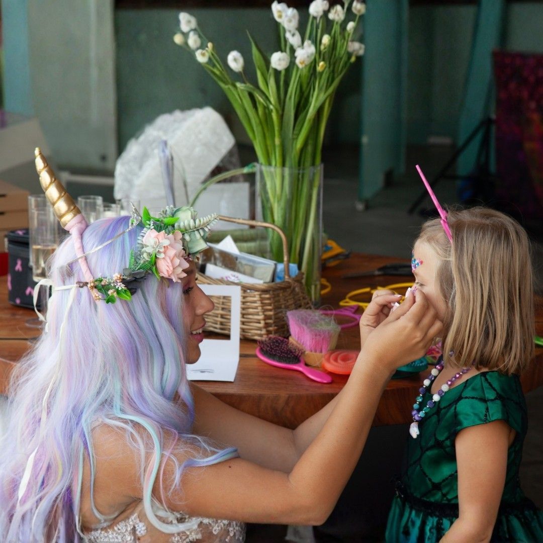 Two children in costumes playing at a table, one painting the other's face near flowers and snacks.