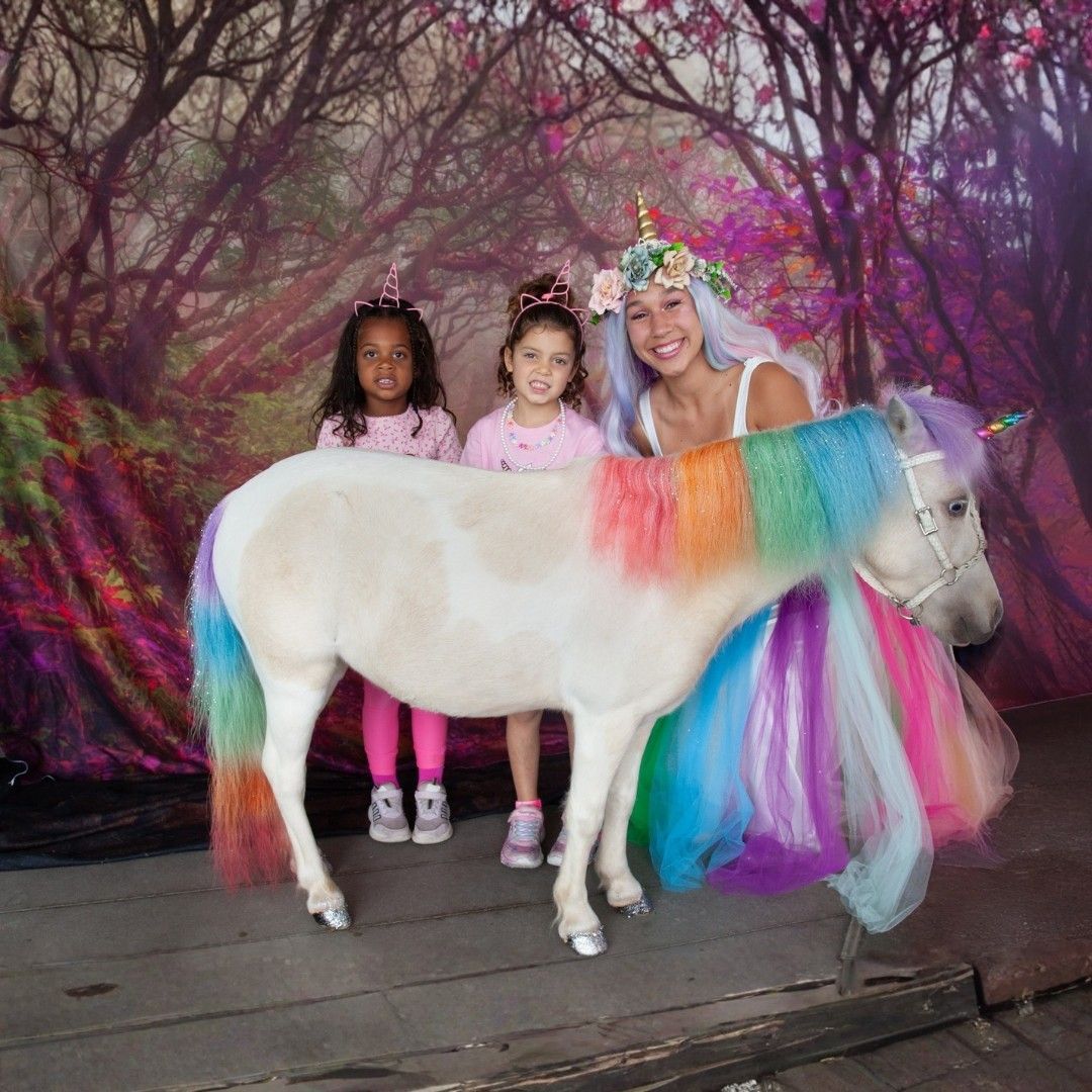 Person in a rainbow tutu posing with two children beside a white unicorn at a whimsical backdrop
