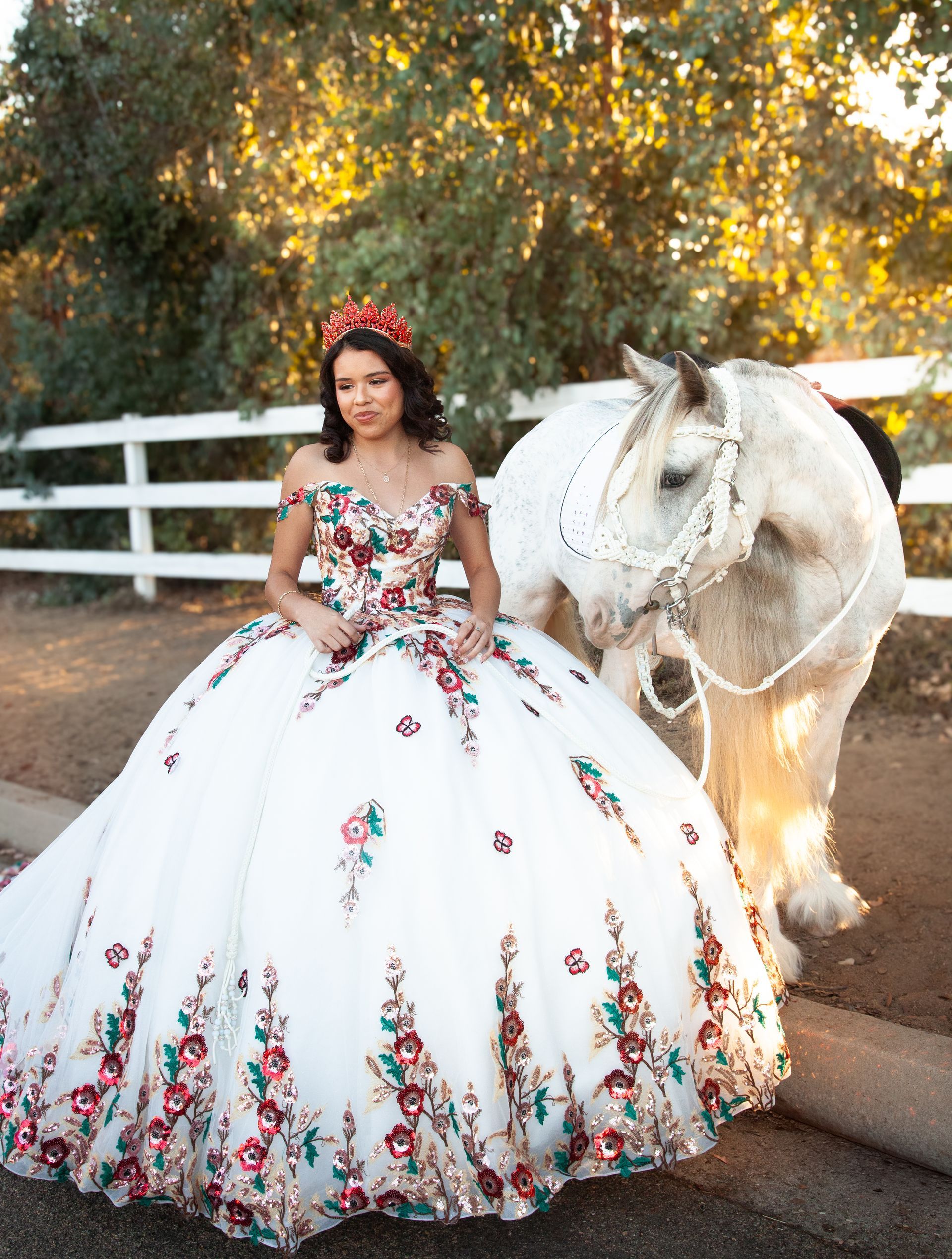 Woman in a floral quinceanera ball gown standing beside a white horse outdoors at sunset for a photoshoot in San Diego