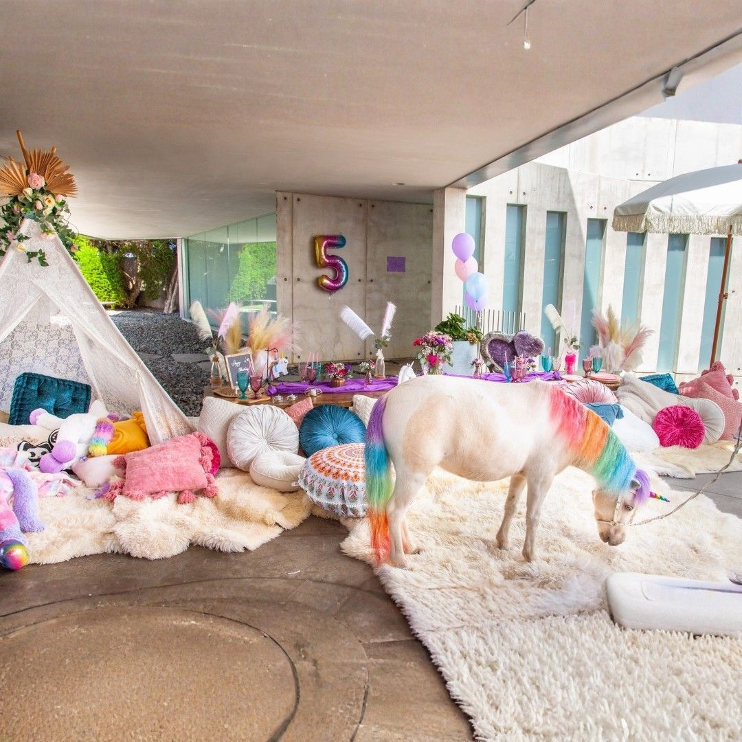 Colorful unicorn-themed party setup with balloons, plush decor, and a large rainbow horse in a bright patio space