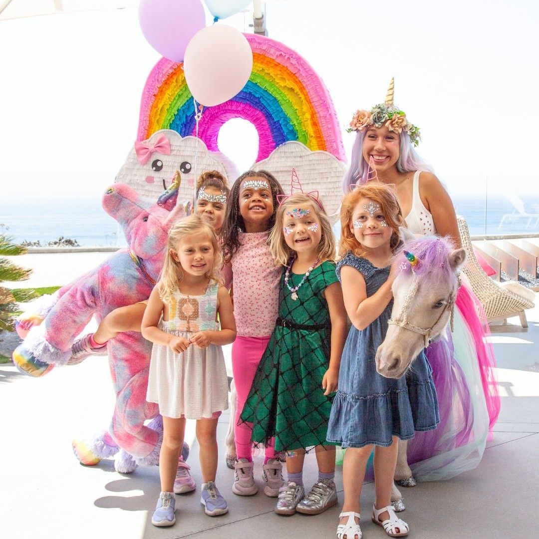 Children posing with a unicorn float and rainbow backdrop at a beach party, smiling in colorful dresses