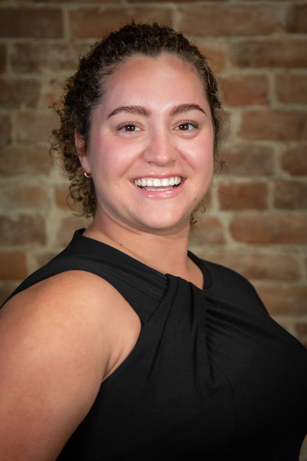 Woman with curly brown hair smiles in a black sleeveless top, brick background.