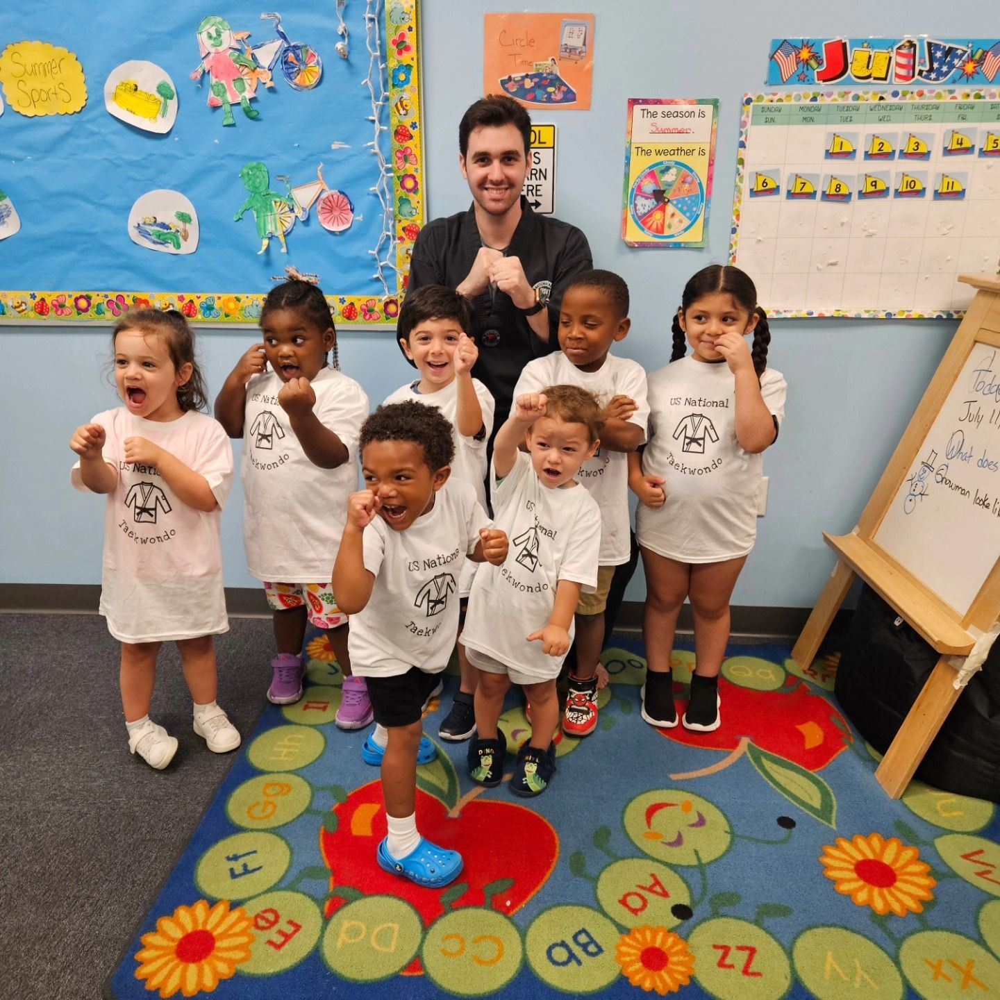 A teacher and seven children in matching white shirts pose with their fists raised in a colorful preschool classroom.