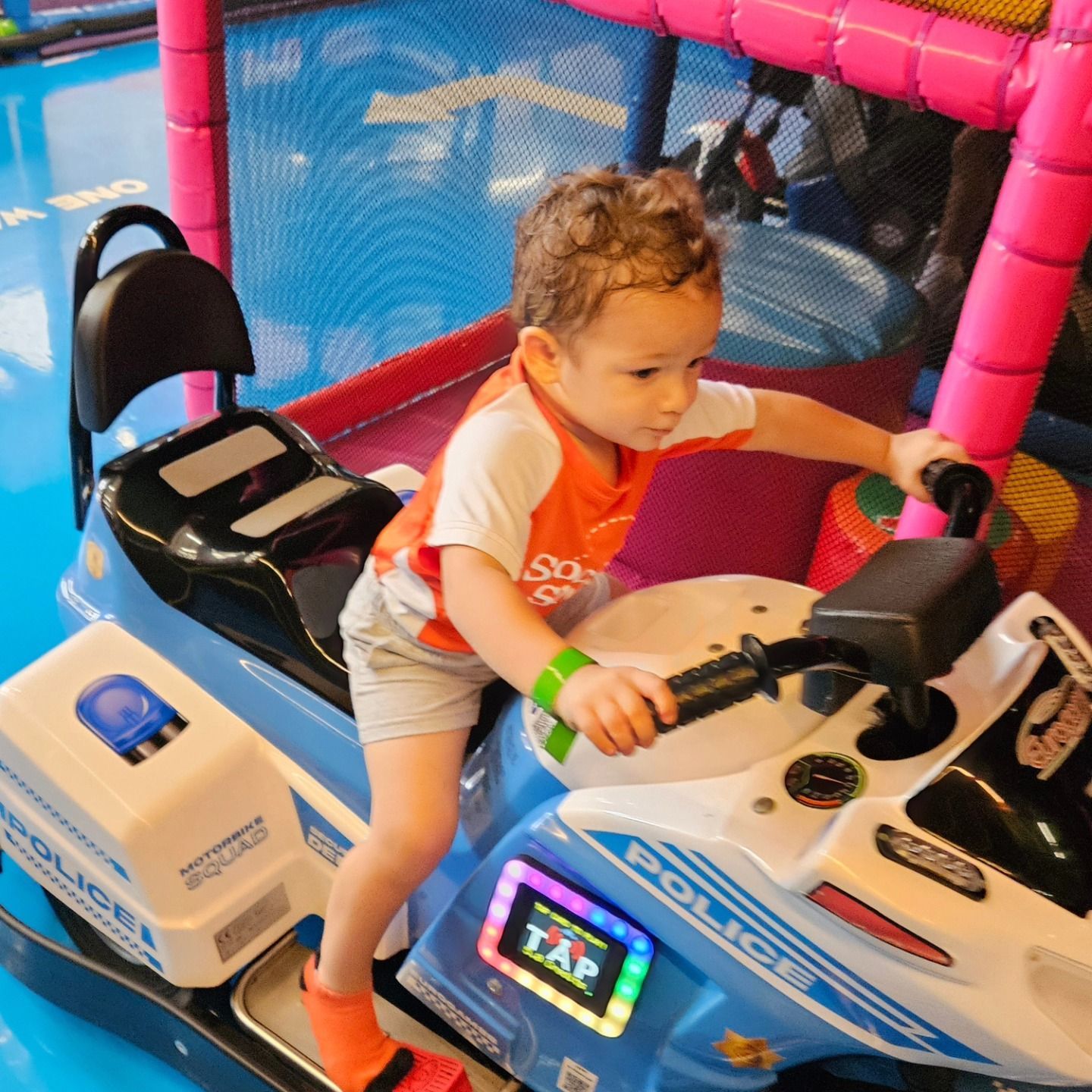 A child in an orange shirt sits on a blue police motorcycle ride-on toy inside a brightly colored indoor play area.