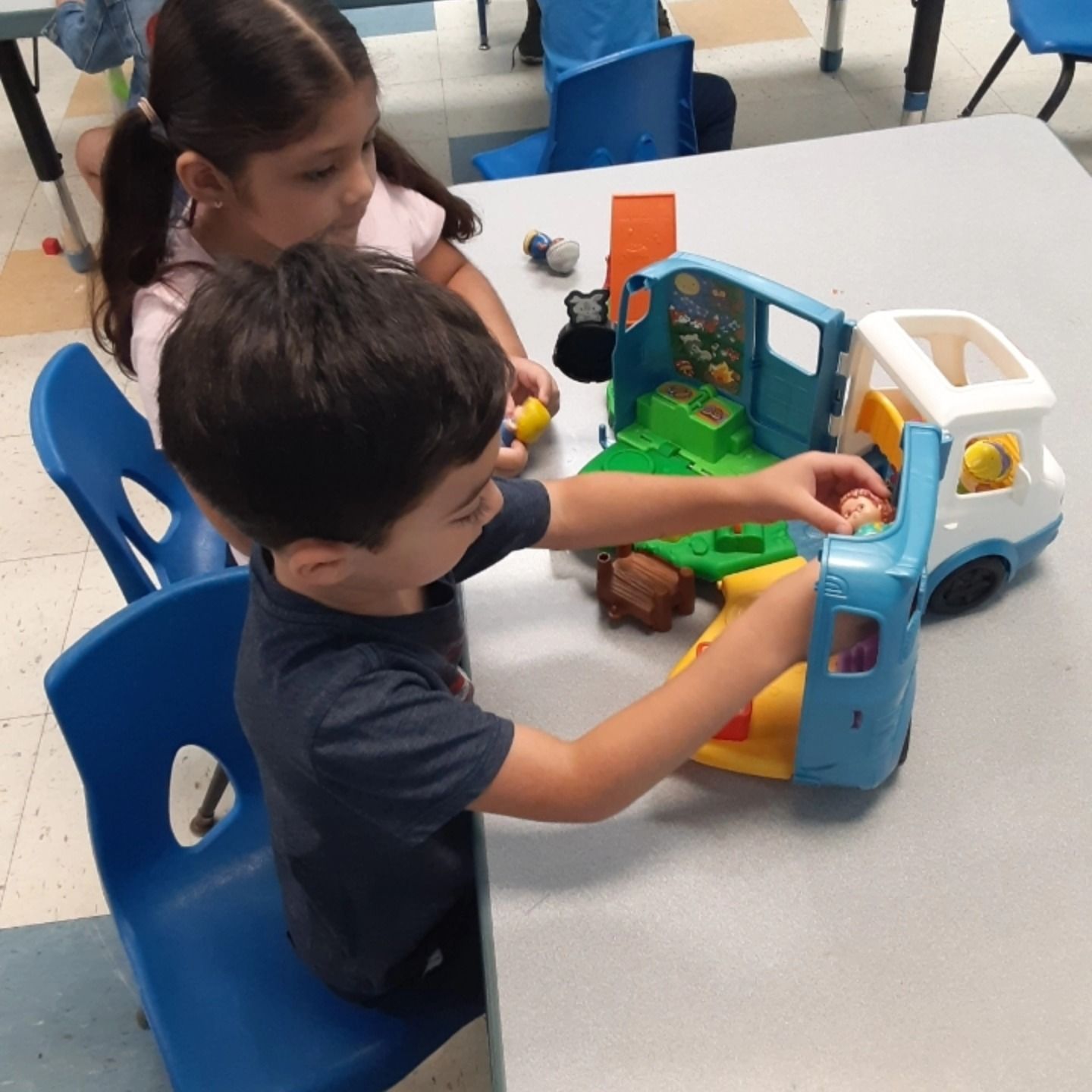 Two children sit at a table playing with a blue and white toy camper set.
