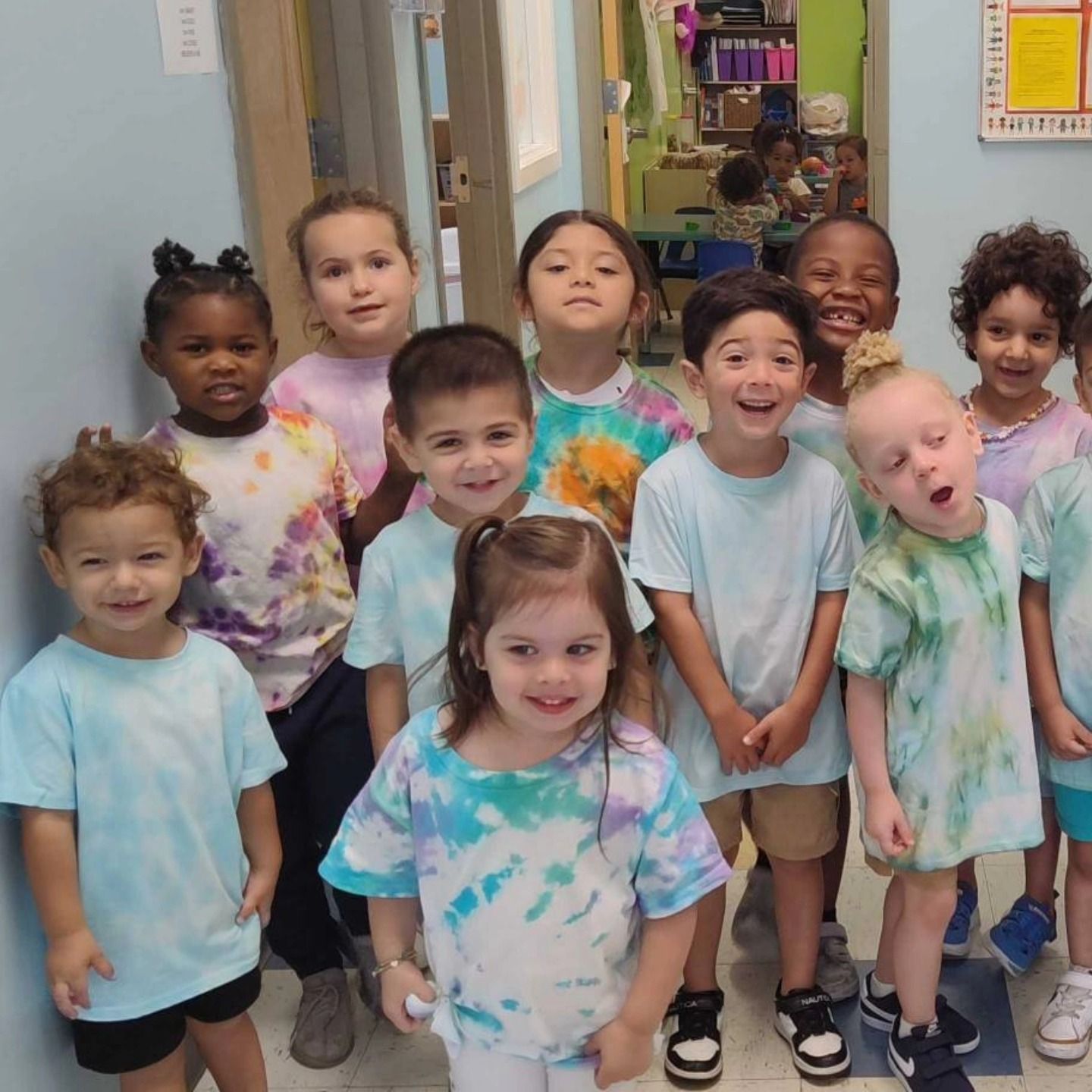 A group of children stand together indoors, smiling while wearing matching tie-dye shirts.