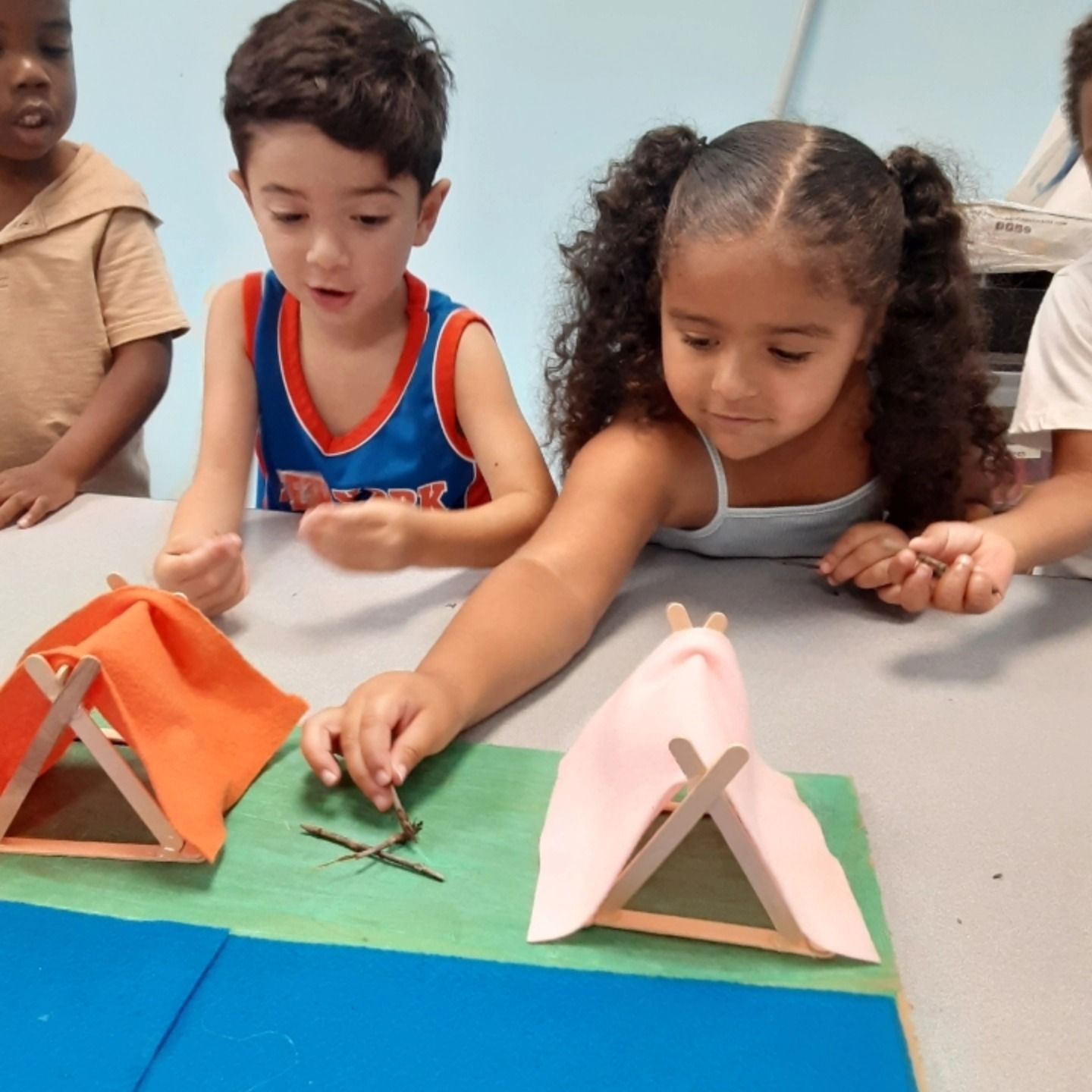 Three children at a table build miniature tents from craft sticks and fabric, one child places sticks on a green mat.
