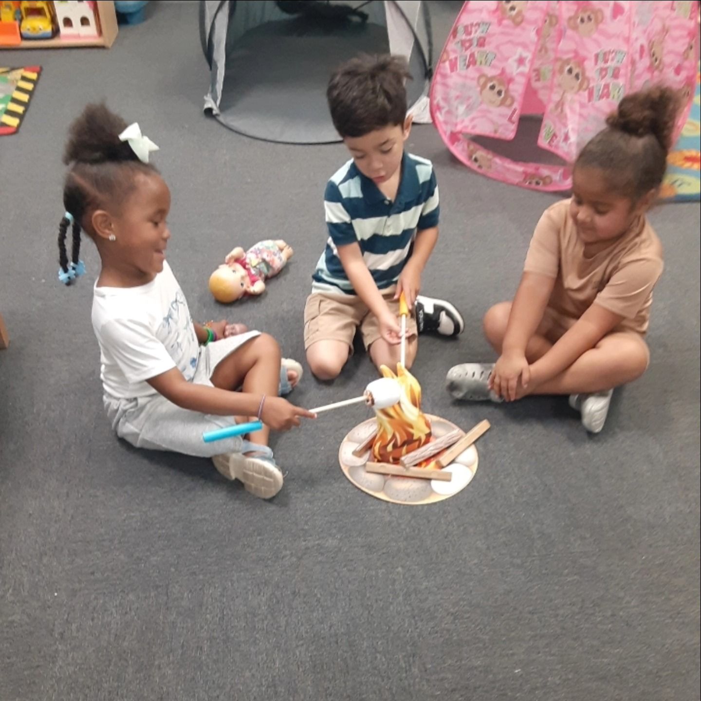 Three children sit in a circle on a gray floor, engaging in a pretend campfire activity with toy flames and sticks.