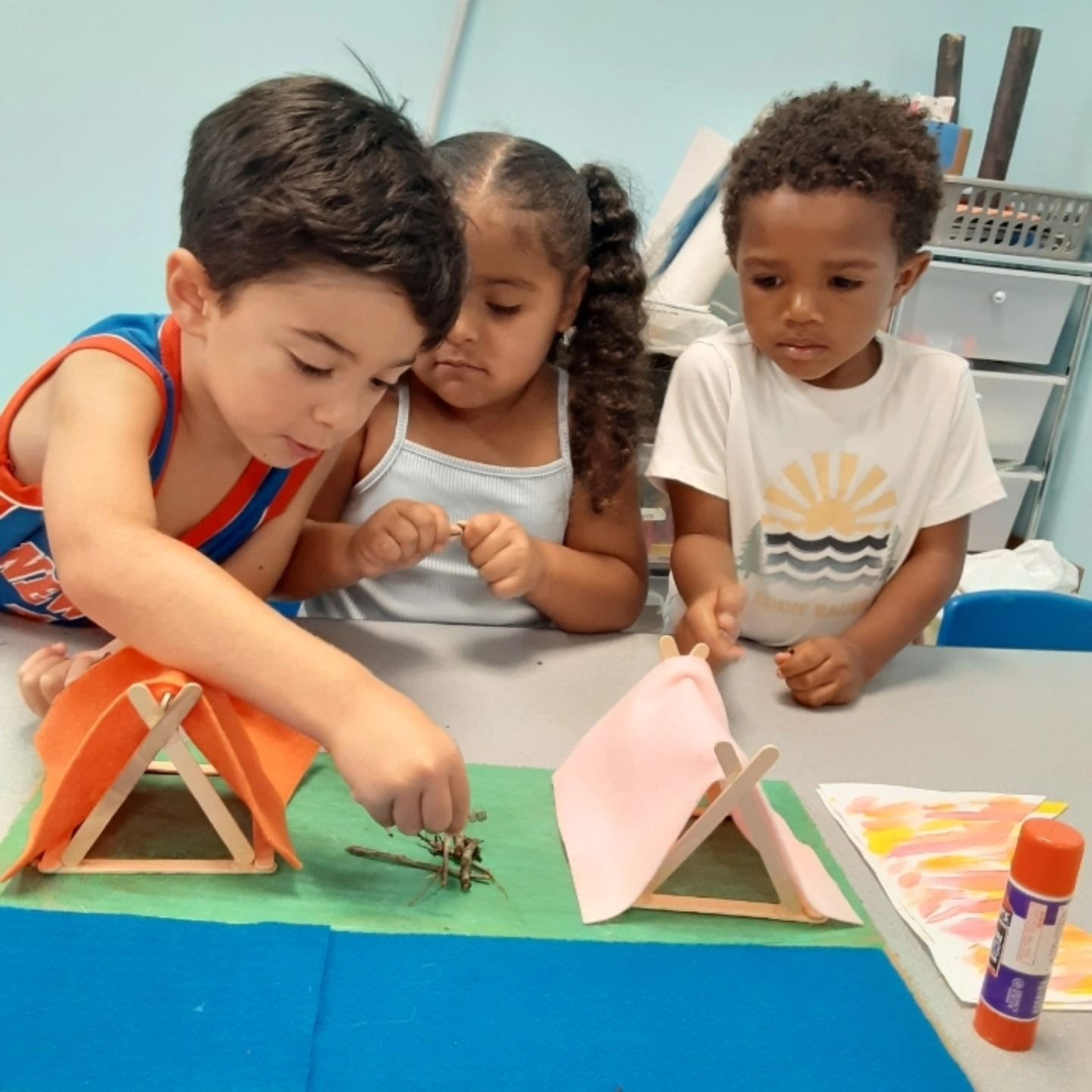 Three children craft a small campsite scene with model tents and twigs on a tabletop.