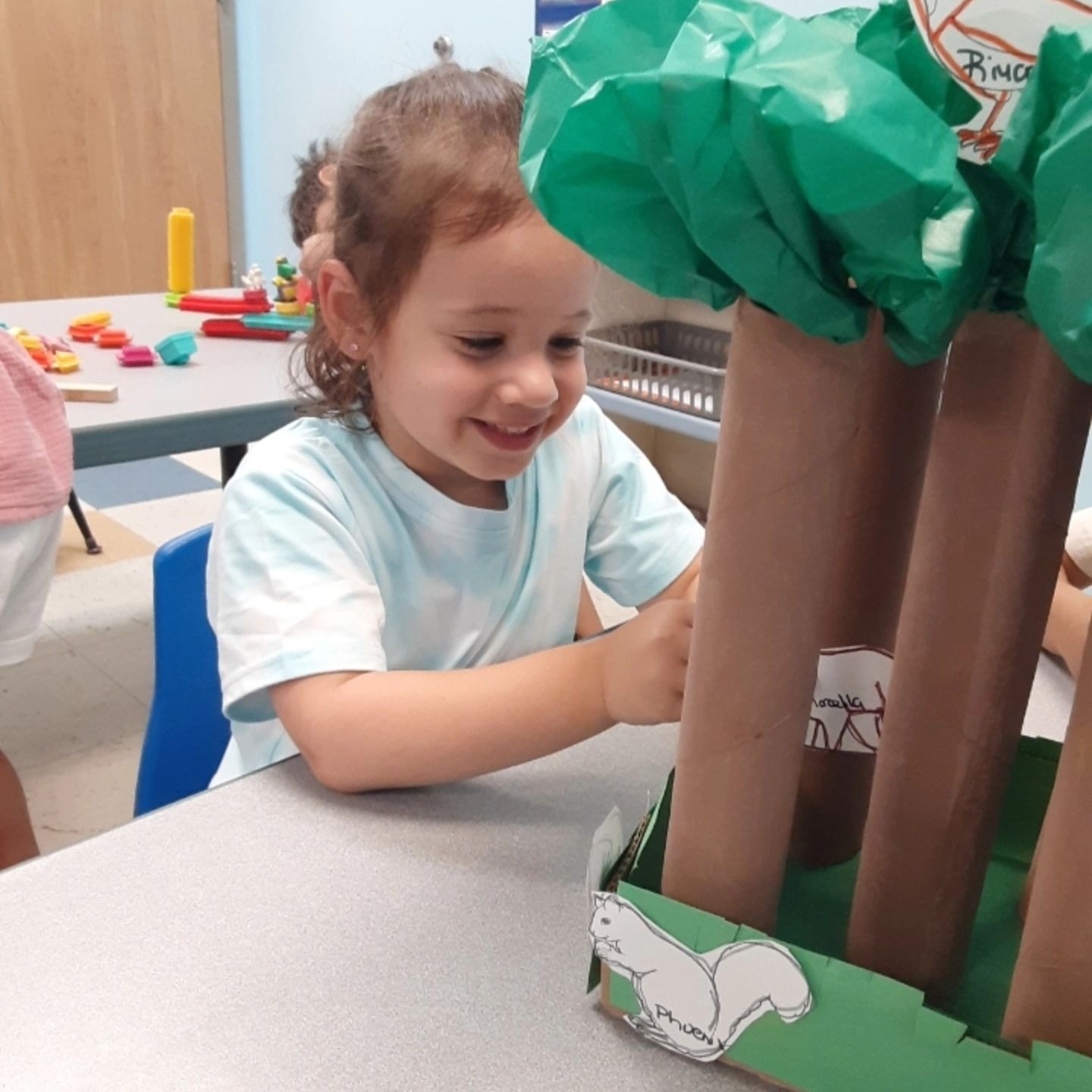 A smiling child interacts with a craft model of a forest made from brown cardboard tubes and green tissue paper.