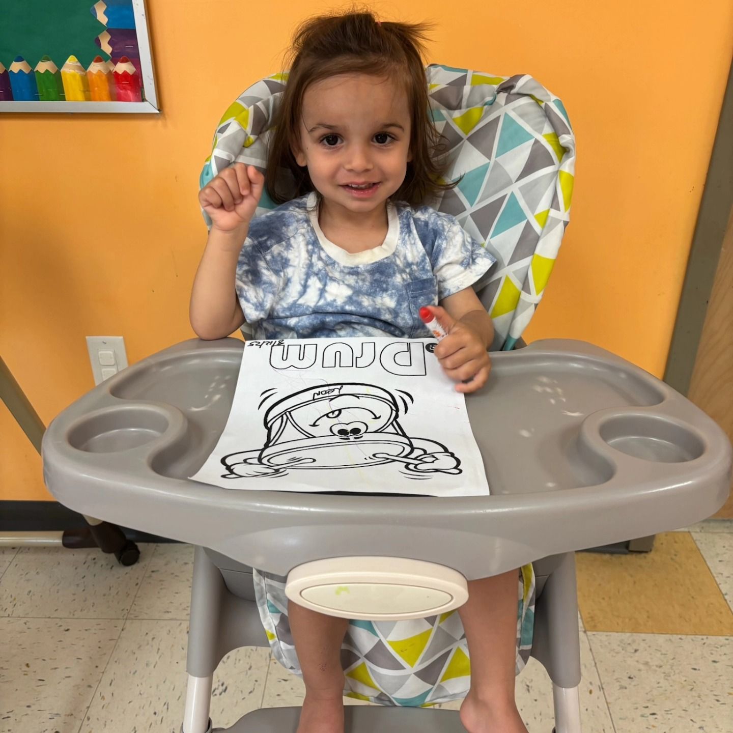 A person sits in a high chair holding a marker, with a coloring sheet featuring the word