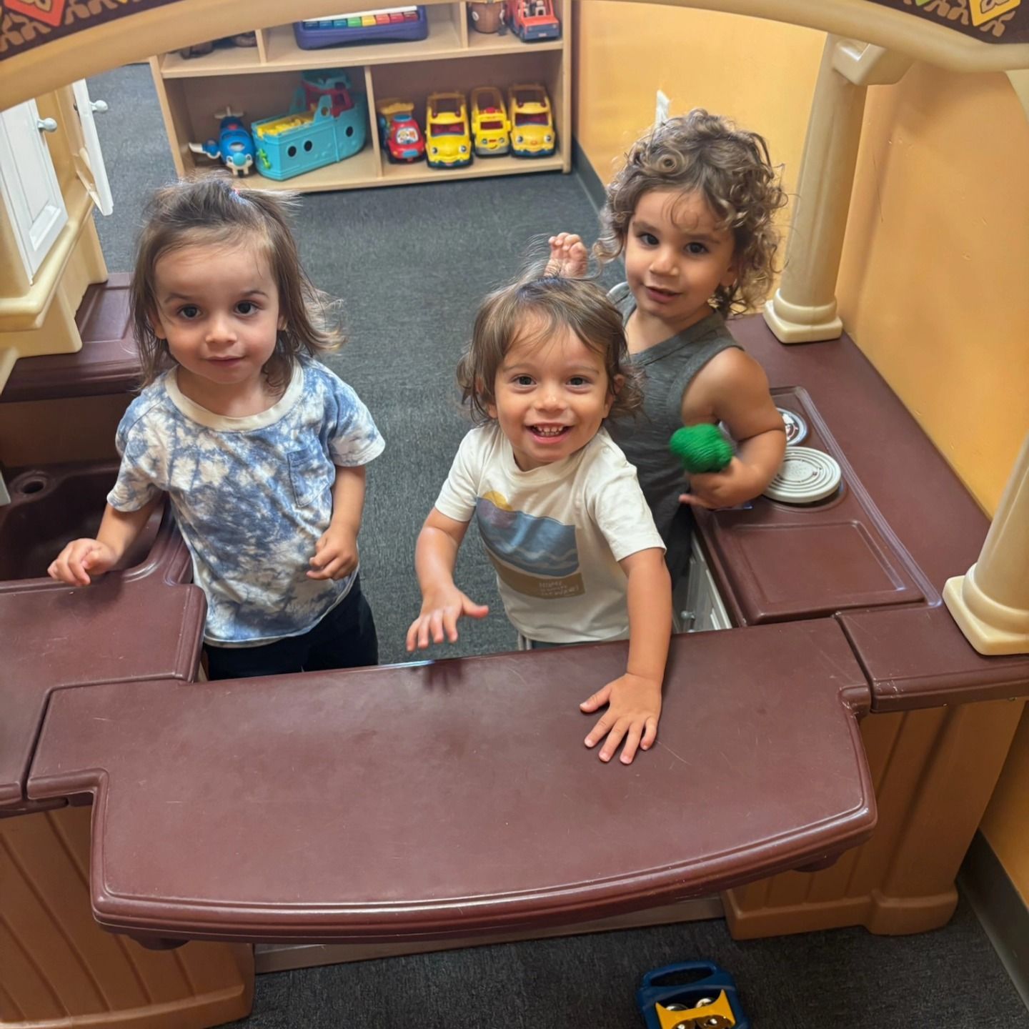 Three children smile while playing together in a toy kitchen inside a playroom.