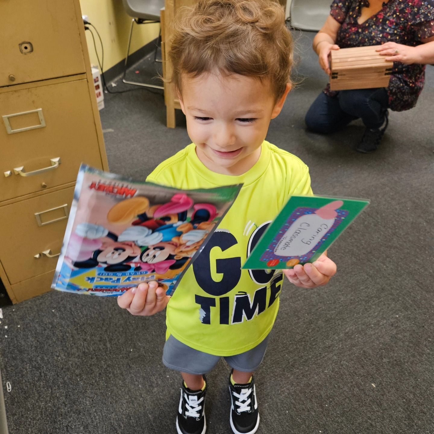 A smiling child in a bright yellow shirt holds a Mickey Mouse activity pack and a small card in an indoor setting.