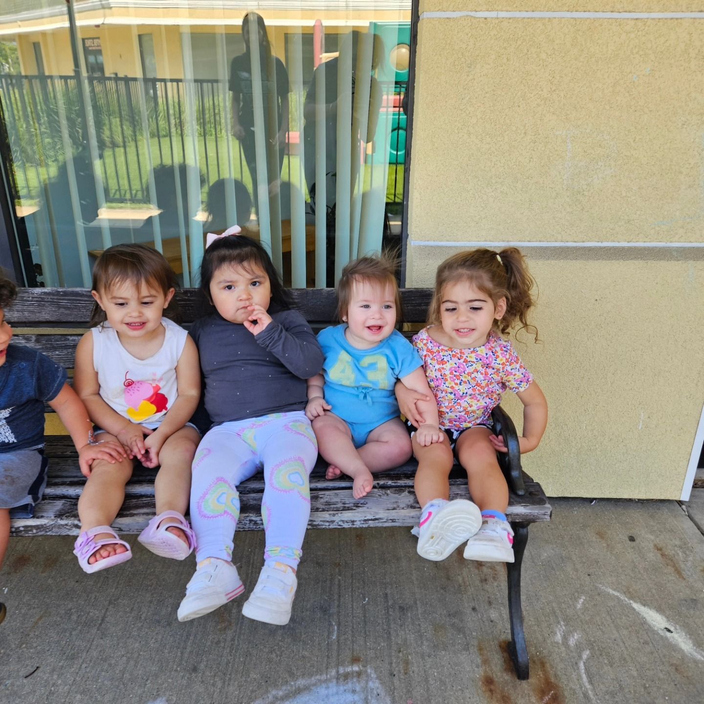 Four children sit together on a weathered wooden bench outdoors, smiling and looking toward the camera.