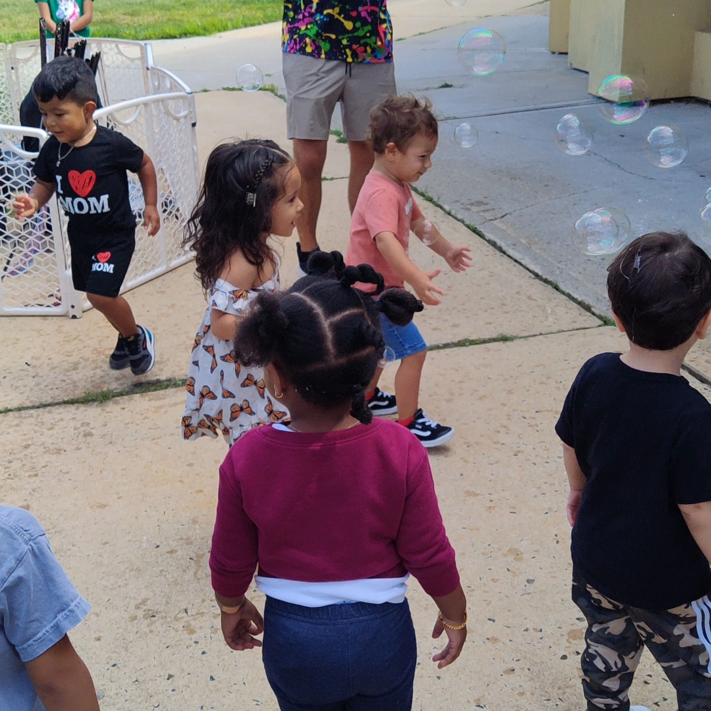 Several children play with bubbles on a paved outdoor area, with an adult partially visible in the background.