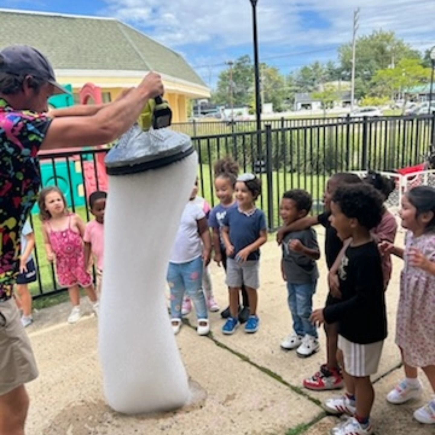 An adult uses a bubble machine to create a tall, vertical stream of foam for a group of children on a fenced playground.