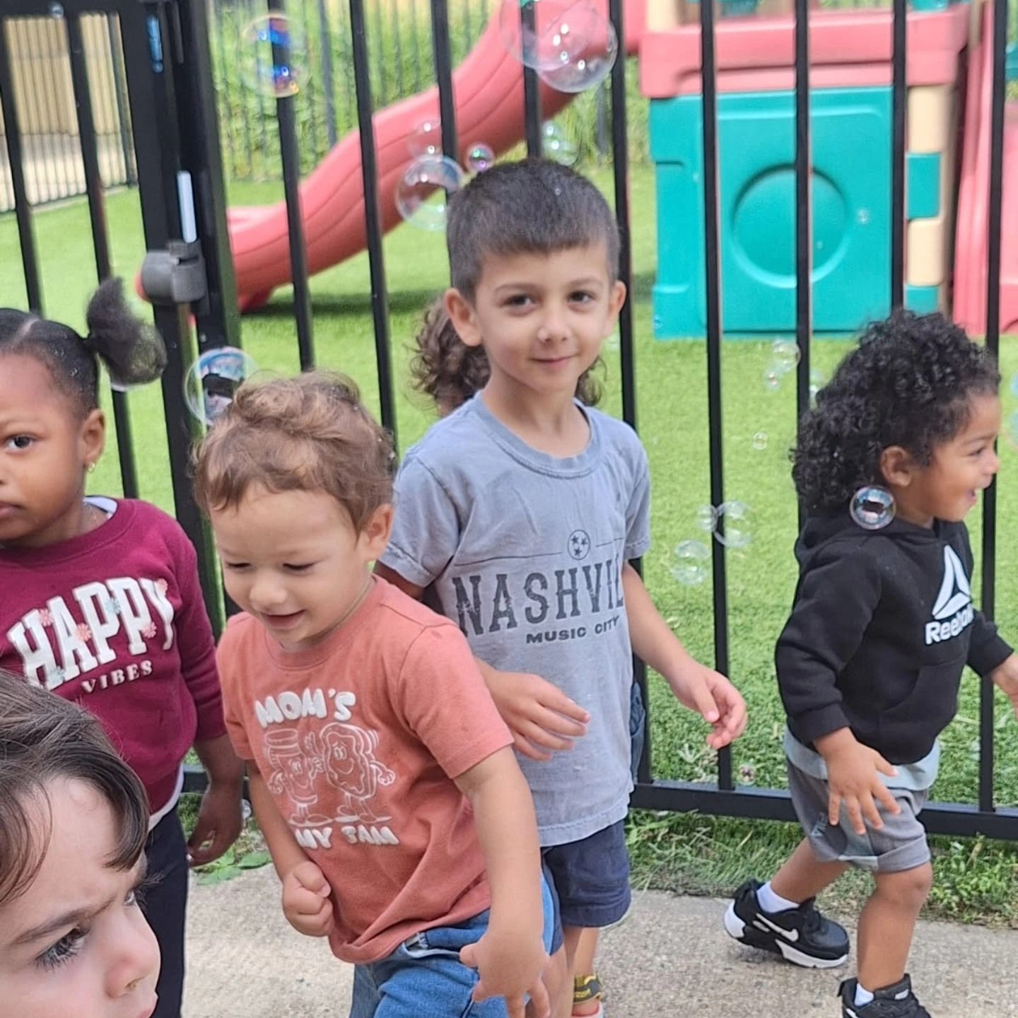 Children play outdoors near a black fence, with bubbles floating around them and playground equipment in the background.