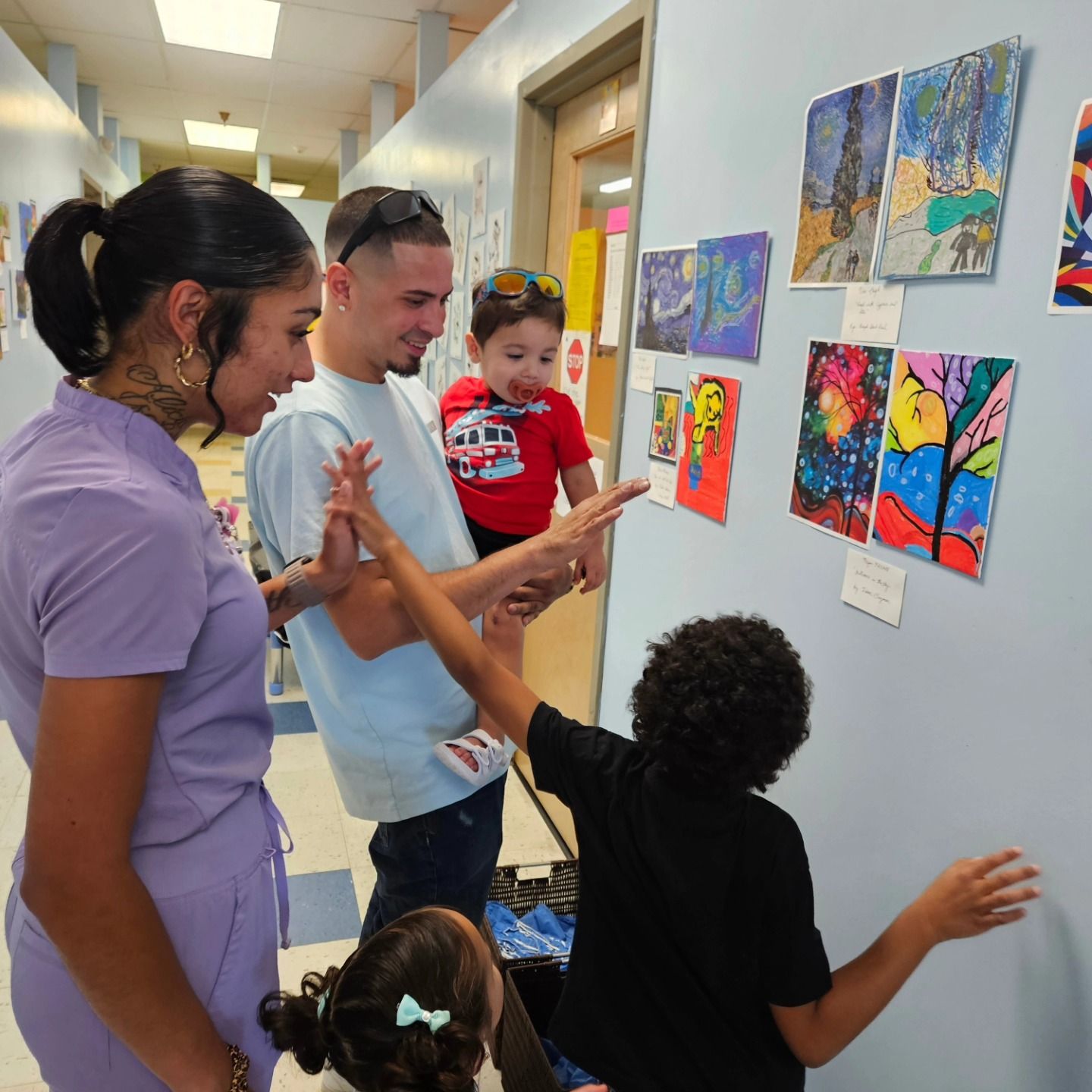 A family looks at colorful children’s artwork displayed on a light blue wall in a school or community hallway.