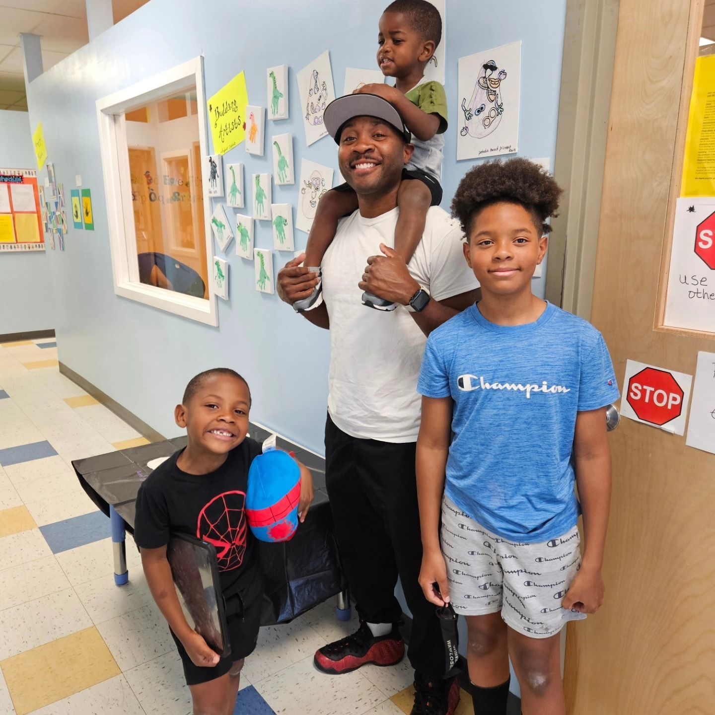 A smiling adult carries a child on their shoulders while two other children stand nearby in a school hallway.