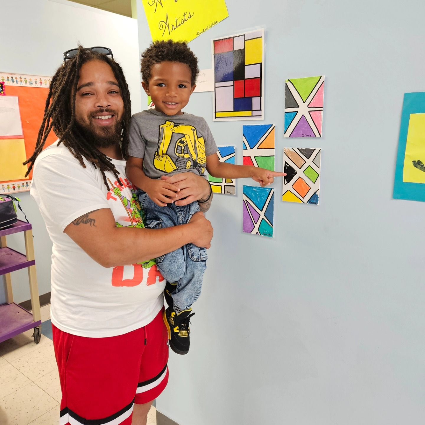 A smiling adult holding a child who is pointing at abstract colorful artwork displayed on a light blue wall.
