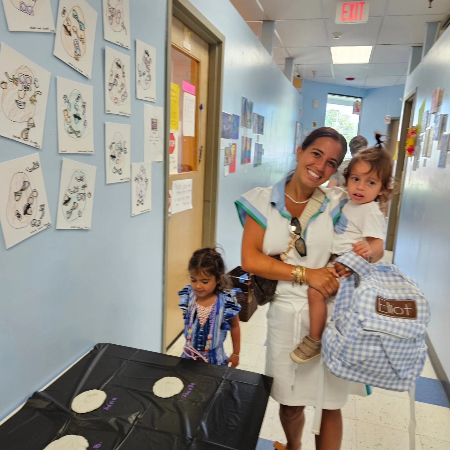 A smiling person carries a toddler in a school hallway with wall art while another child stands nearby.