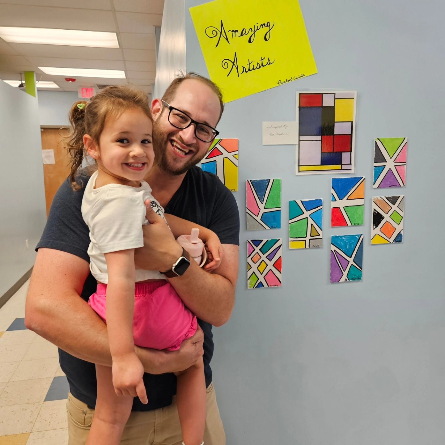 A smiling man holding a child in front of a wall displaying children's art projects under a sign reading