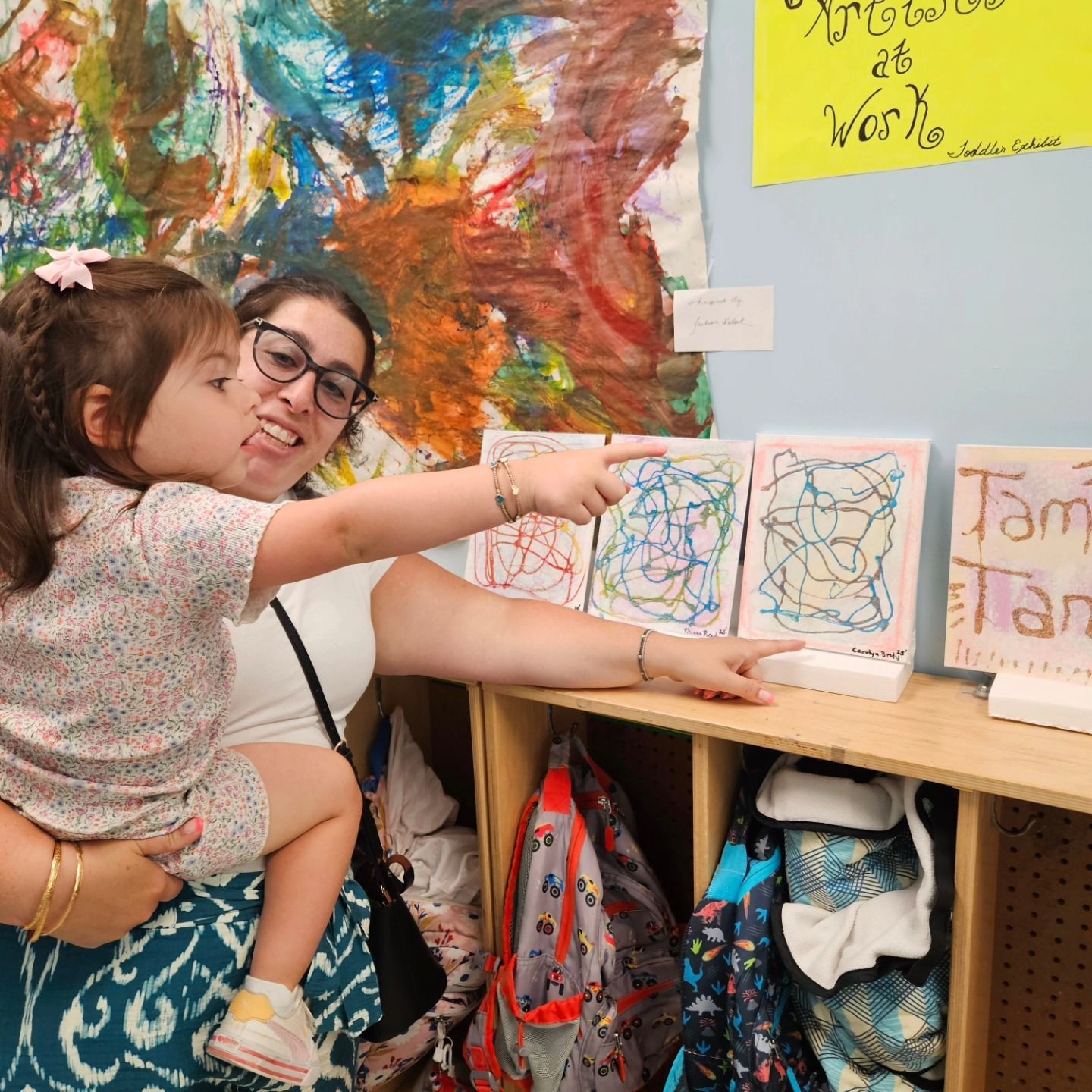 A person holding a child, both pointing at children’s artwork displayed on a wooden shelf.
