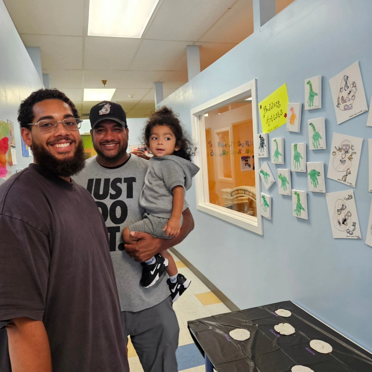 Two adults and a toddler stand in a school hallway near a wall decorated with children’s artwork.
