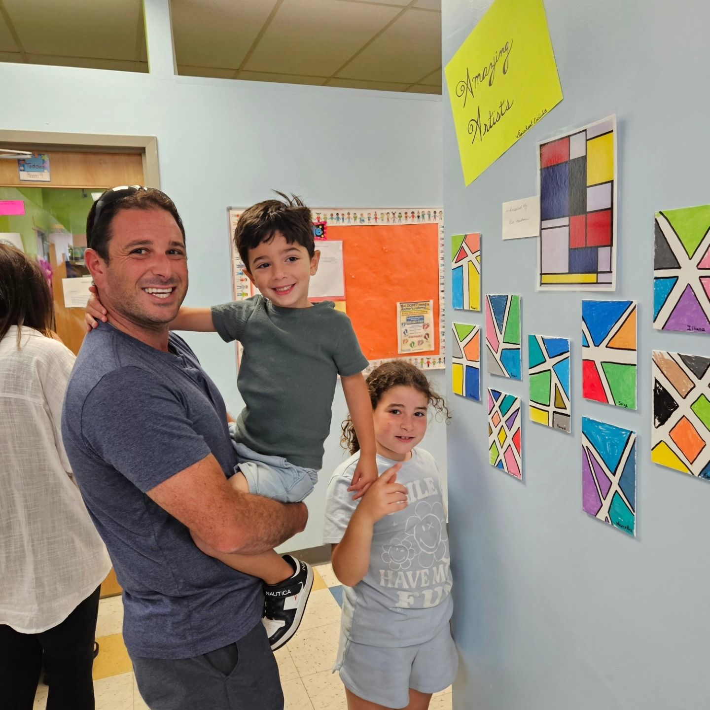 A parent holds a child next to a younger child, all smiling in front of a wall of colorful geometric student artwork.