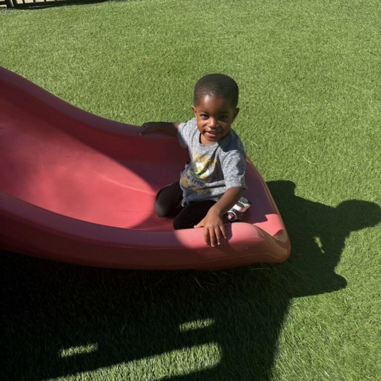 A child in a grey t-shirt sits on a red plastic playground slide on a green turf field on a sunny day.