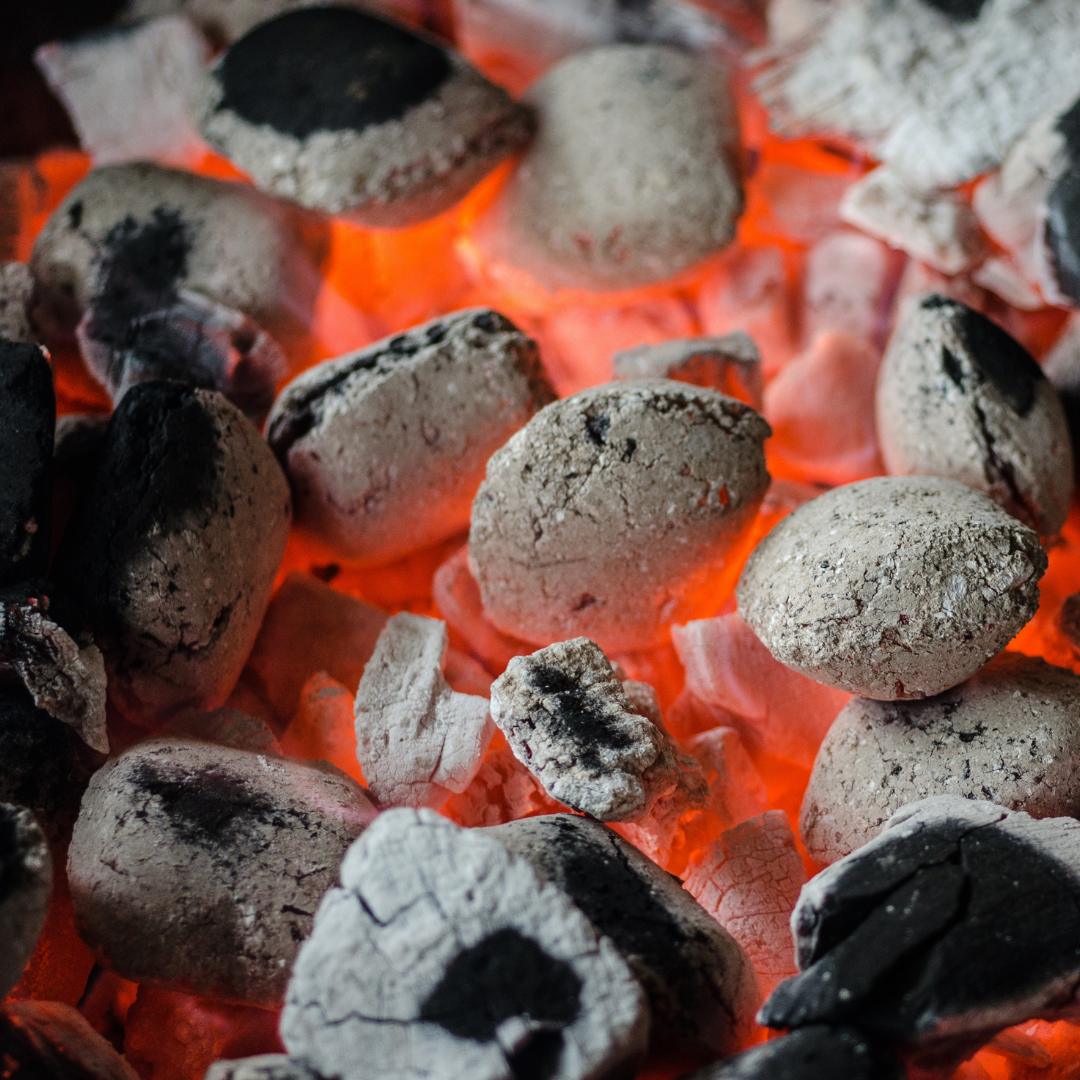 Glowing charcoal briquettes, close up. Red-orange embers visible beneath. Some briquettes are black, some are gray.