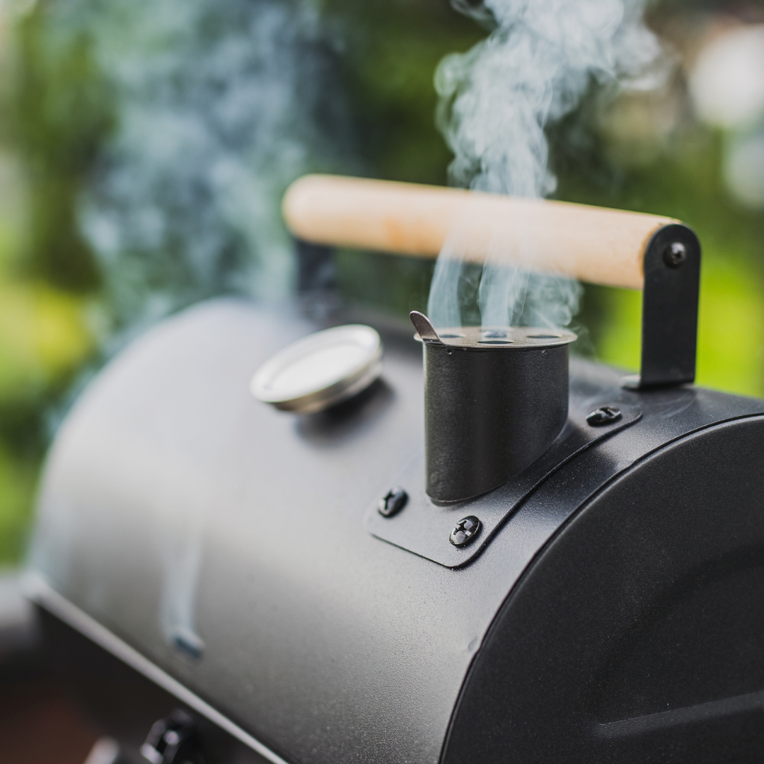 Black smoker grill with smoke rising, wooden handle, thermometer, outdoor setting.