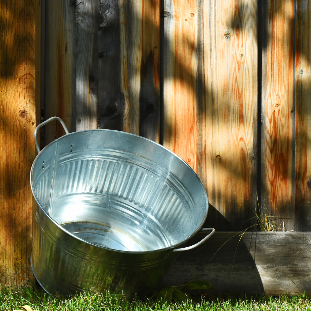 Galvanized metal tub on grass, leaning against a weathered wooden fence.