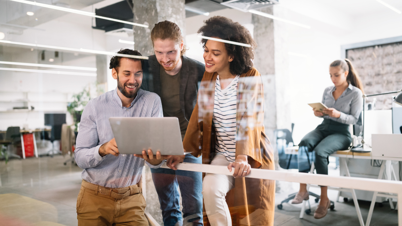 A group of people are standing in an office looking at a laptop.