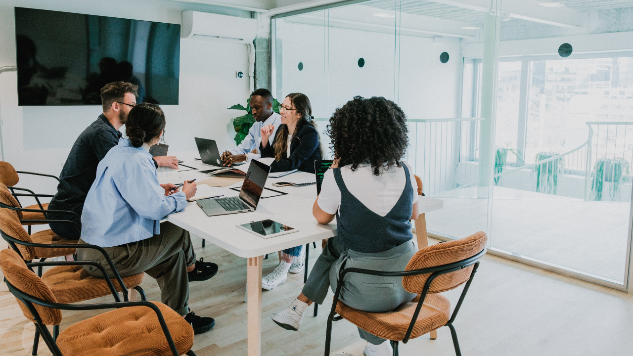 A group of people are sitting around a table in a conference room with laptops.