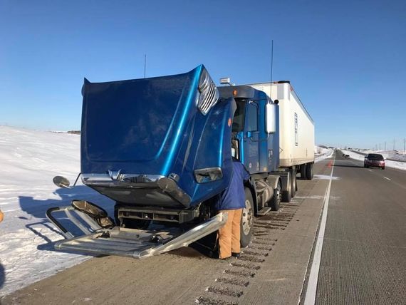 Blue semi-truck with damaged front end parked on snowy roadside beside a highway