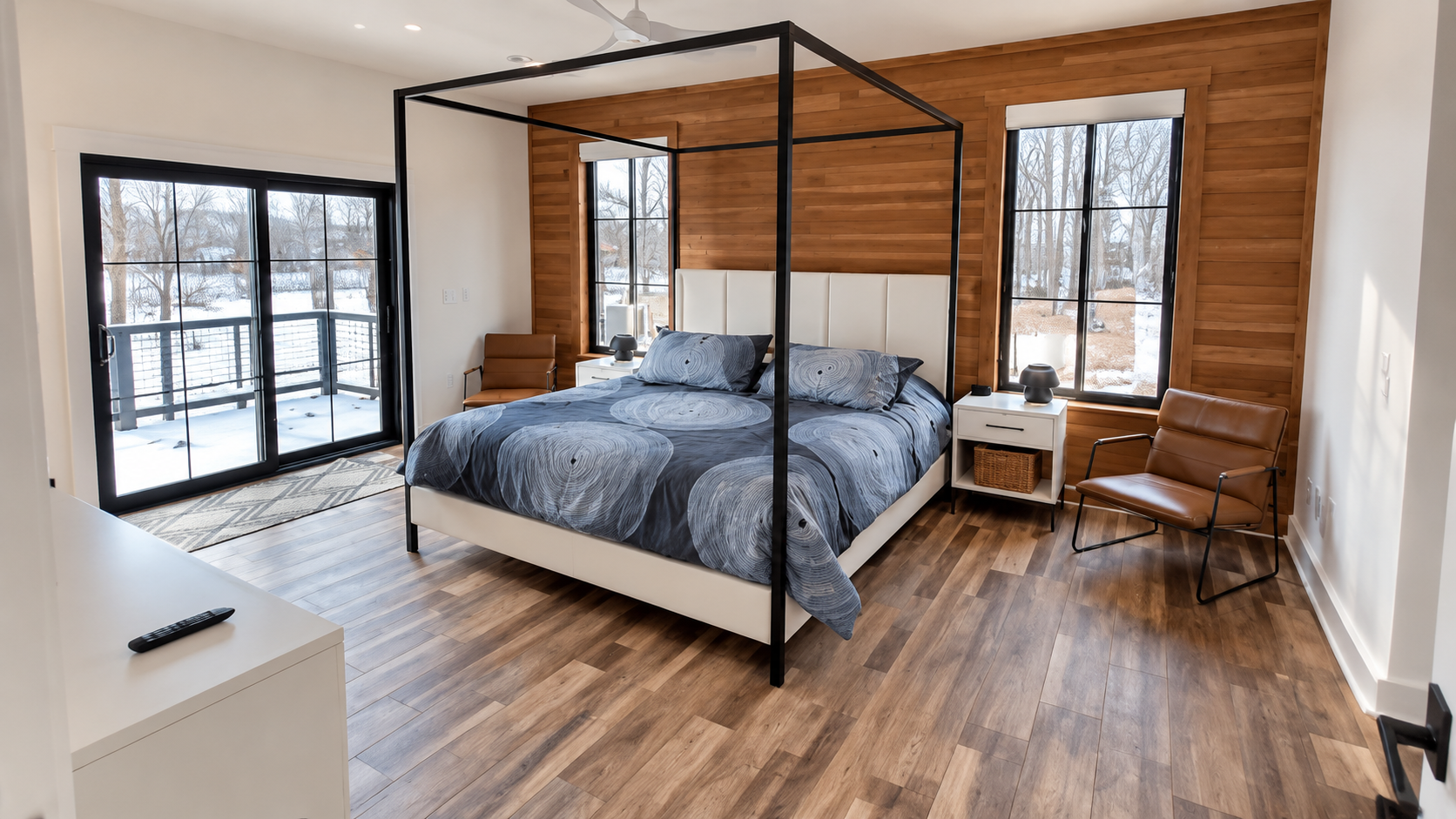 Modern bedroom with black canopy bed, blue bedding, wood floors, and large windows by a small desk