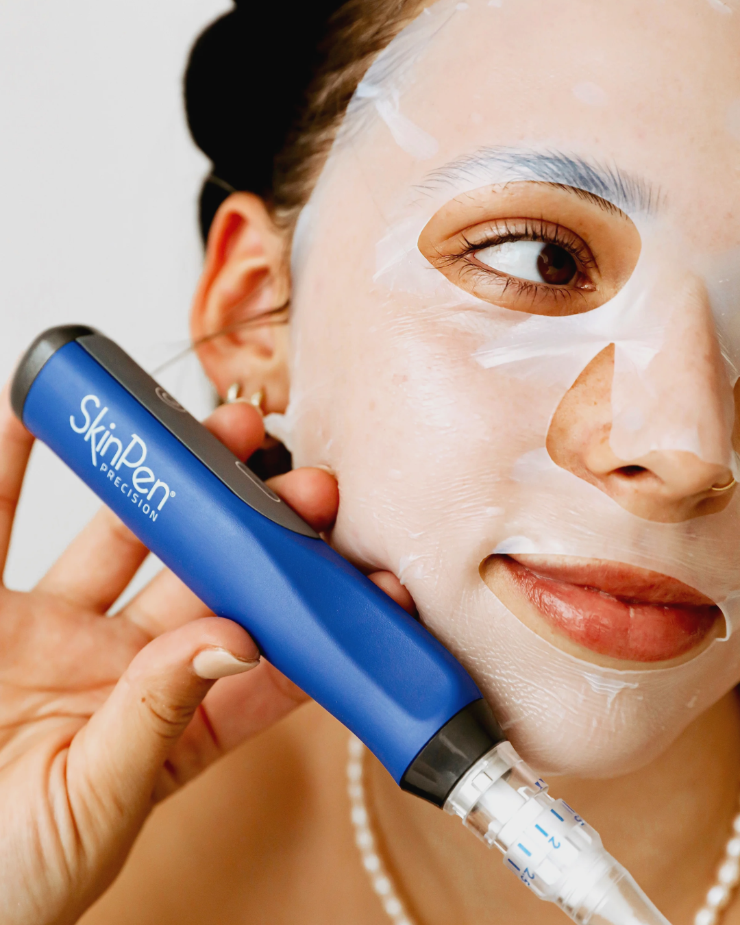 Woman applying facial cream with a blue skincare device, wearing a white mask.