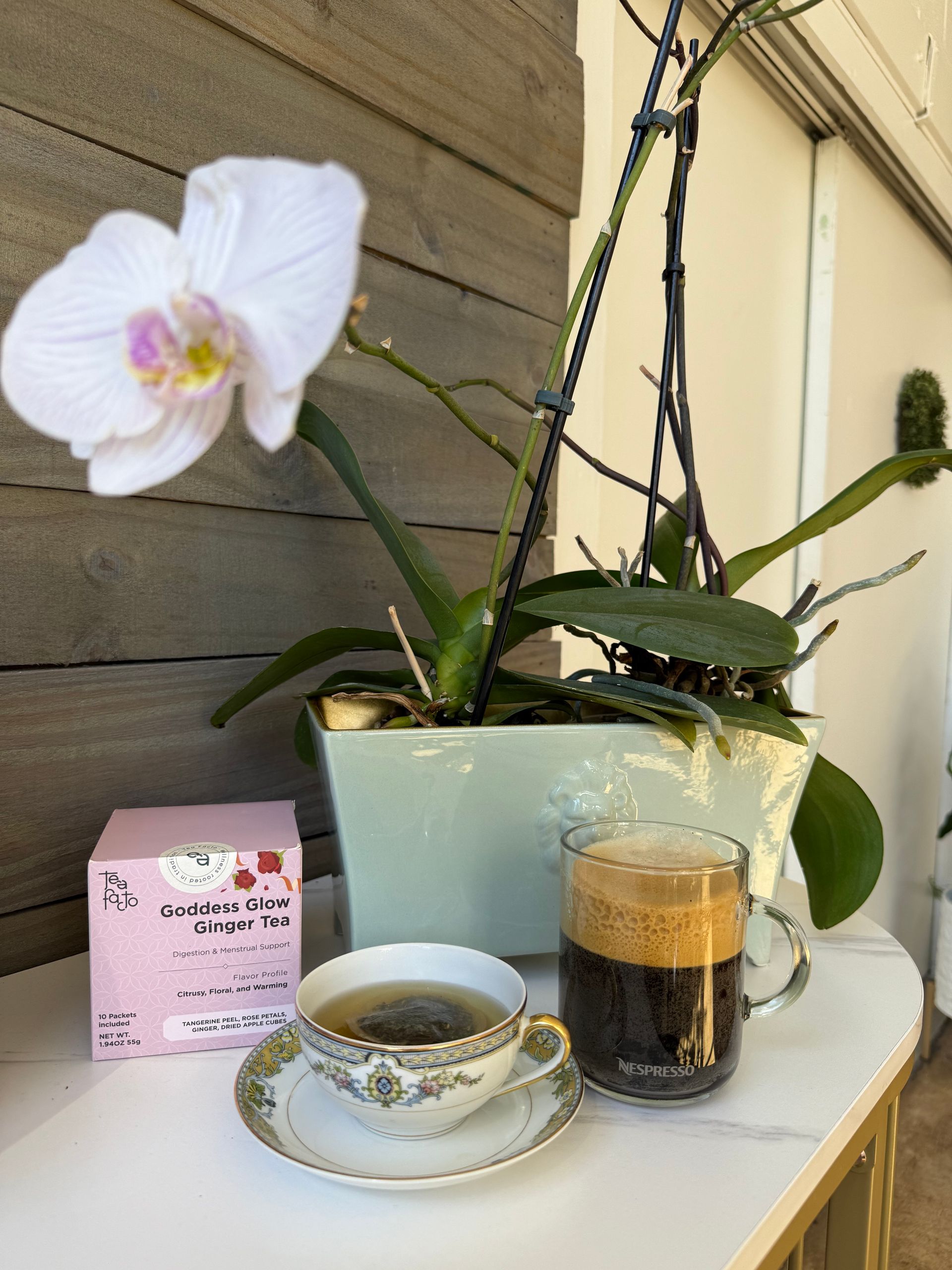 White orchid on a table with teacup, glass drink, and pink box beside a potted plant.