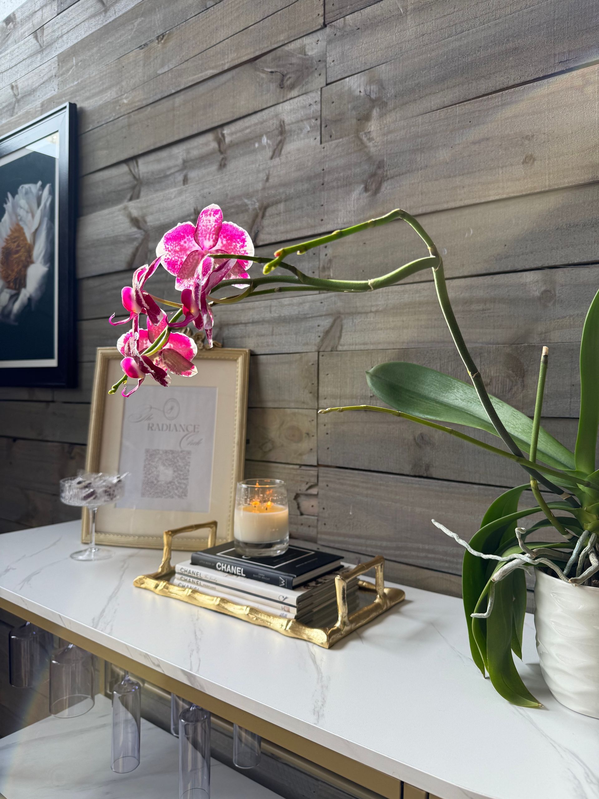 White console table with pink orchid, candle, framed art, and plant against a wood wall.