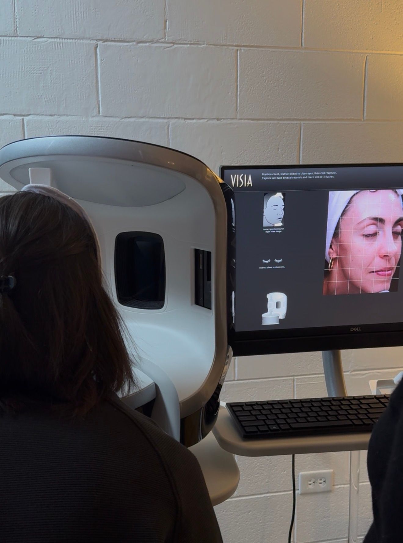 Person seated at a facial imaging station beside a monitor showing a facial scan and close-up face image
