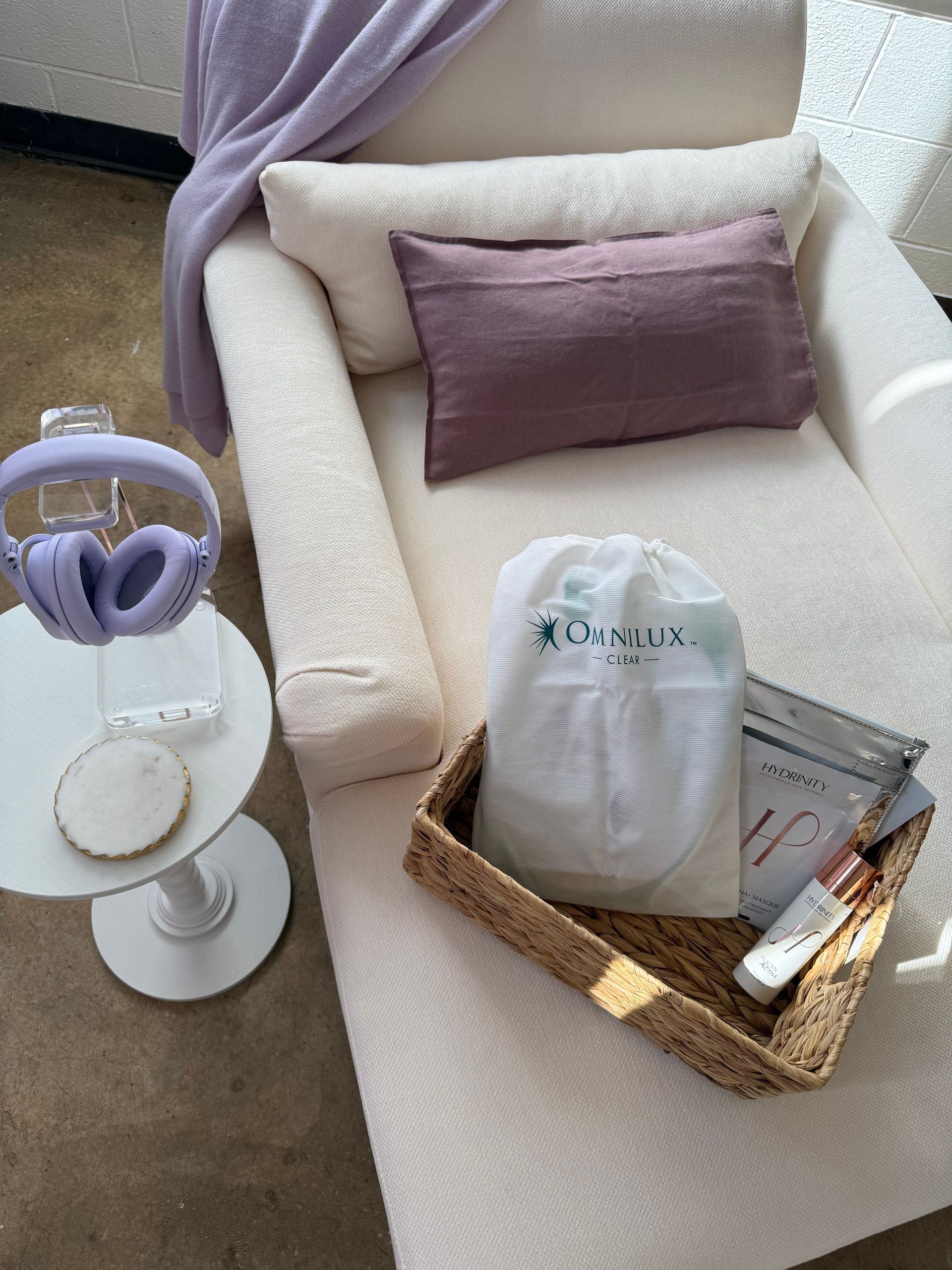 Cream armchair with lavender pillow, side table holding headphones and a drink, and a basket of books and tissues.