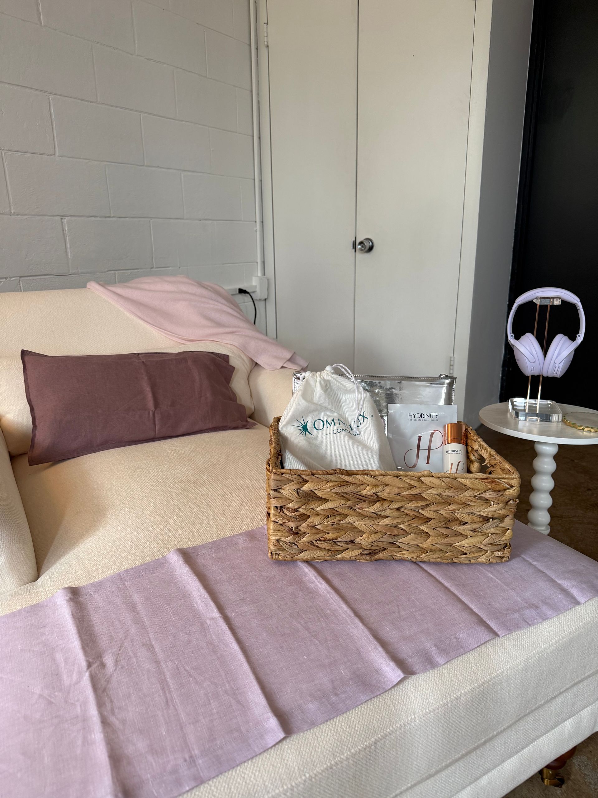 Cozy bed with lavender bedding and pillows, basket of pillows, and side table with headphones in a bedroom corner