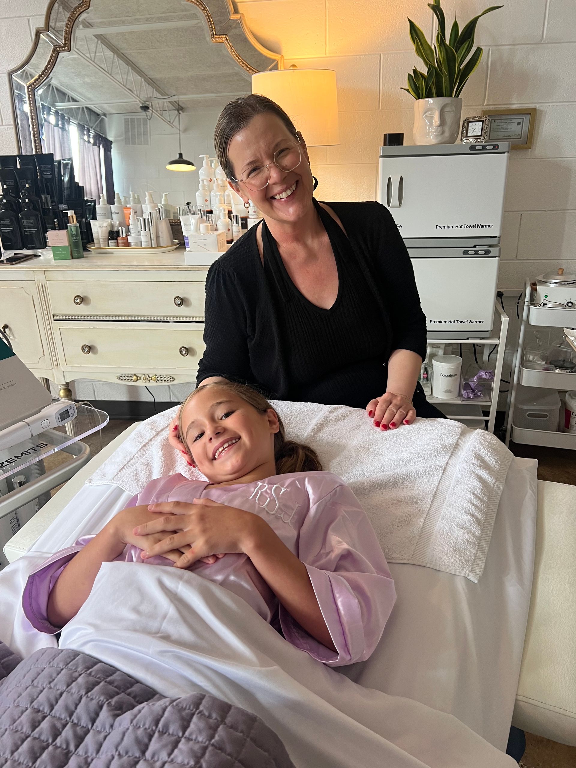 Person smiling beside a reclining client in a bright salon with treatment bed and white cabinets
